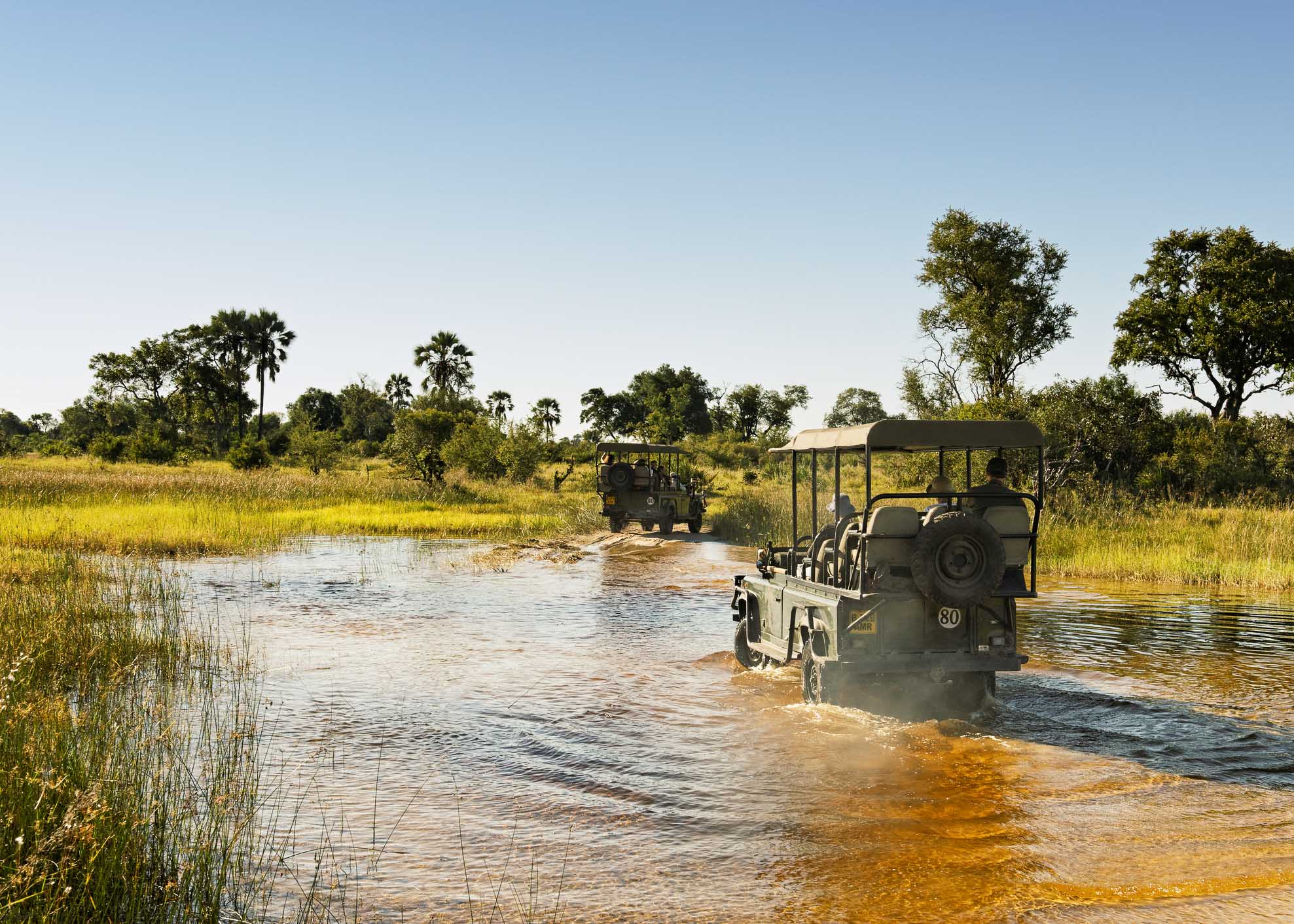 Safari vehicles with tourists driving through the water at the end of the rainy season during a game drive in the Okavango Delta.